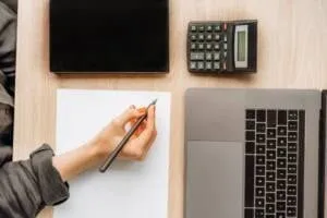um computador uma calculadora e uma pessoa com lápis na mão prestes a escrever