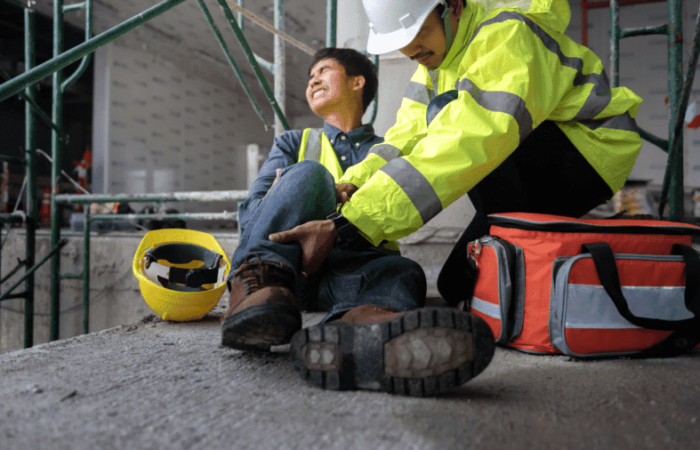 homem caído no chão após uma queda com o pé machucado sendo socorrido por colega de trabalho