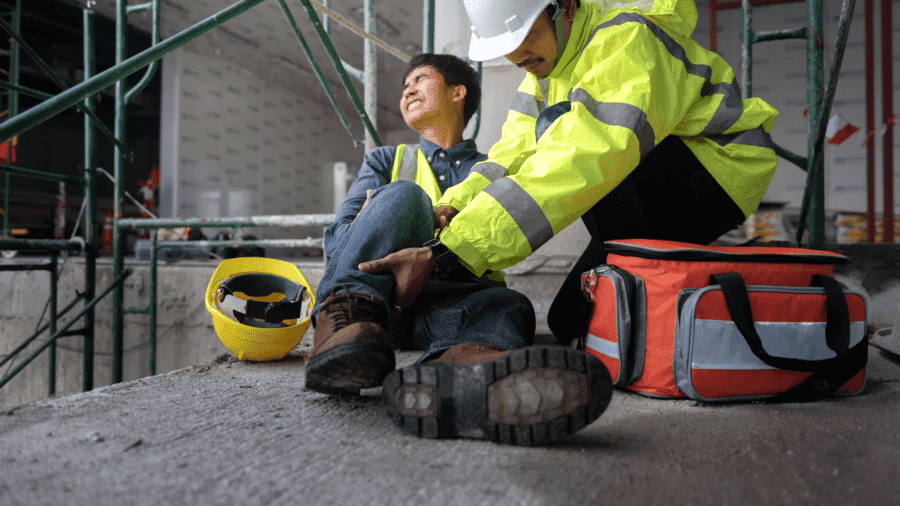 homem caído no chão após uma queda com o pé machucado sendo socorrido por colega de trabalho