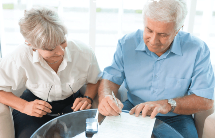 Prova de Vida no INSS 2024 1 homem está assinando papel com a esposa assistindo e ambos sorrindo